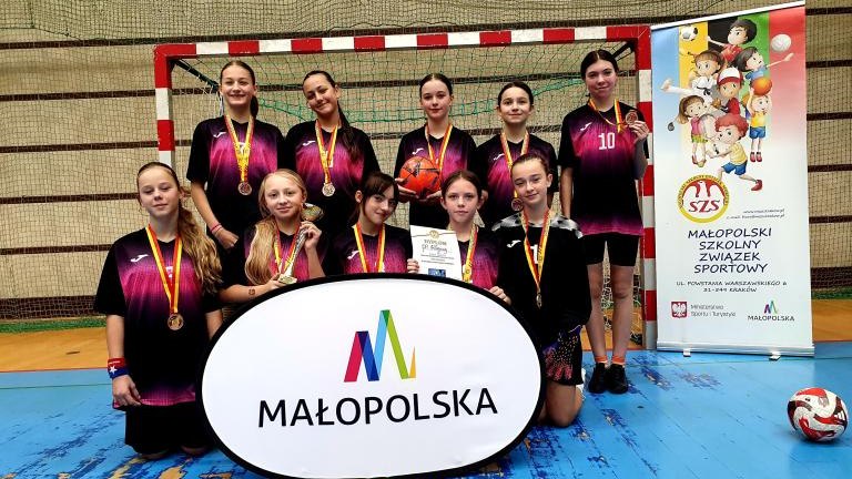 Dziewczęta z SP Maniowy z dyplomem i medalami trzymające plansze z logotypem województwa Małopolskiego na tle brami.jpg